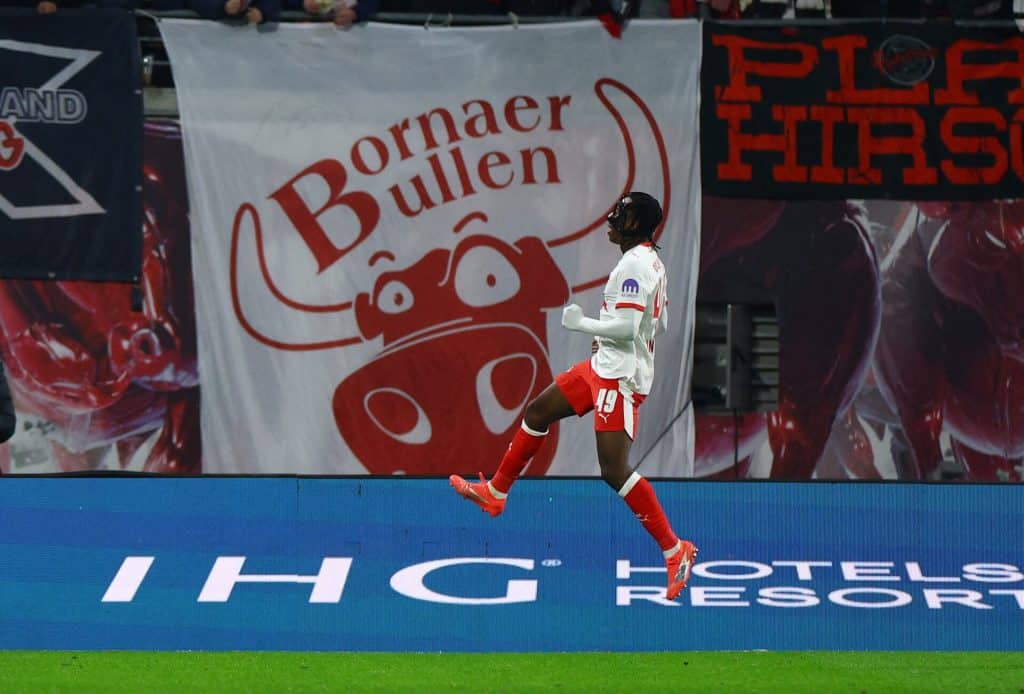 Athletic football player jumping in celebration on the field during a game, with advertising banners in the background.