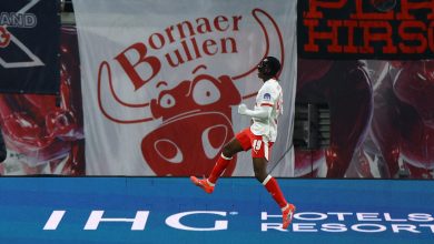 Athletic football player jumping in celebration on the field during a game, with advertising banners in the background.