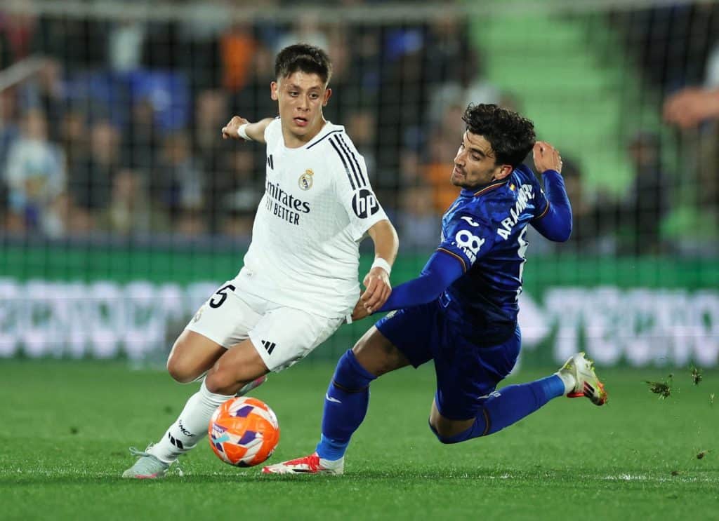 Two young male soccer players in action during a competitive match, one in white jersey and one in blue, battling fiercely for possession on a green field with a stadium background.