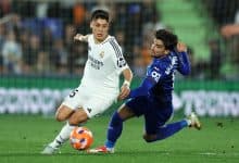 Two young male soccer players in action during a competitive match, one in white jersey and one in blue, battling fiercely for possession on a green field with a stadium background.