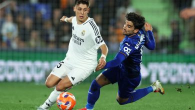 Two young male soccer players in action during a competitive match, one in white jersey and one in blue, battling fiercely for possession on a green field with a stadium background.