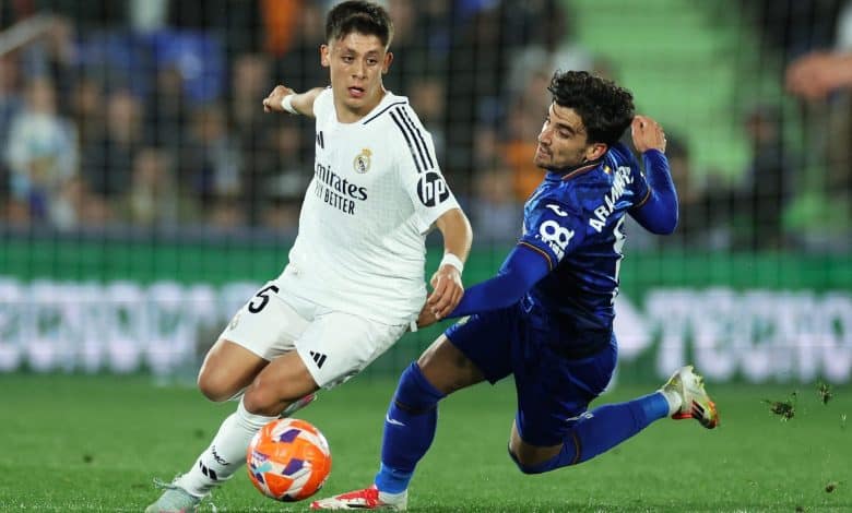 Two young male soccer players in action during a competitive match, one in white jersey and one in blue, battling fiercely for possession on a green field with a stadium background.