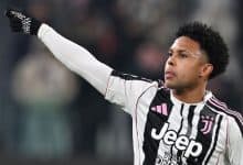 Soccer player in Juventus kit raising arm during match, focused on game, with blurred stadium background.