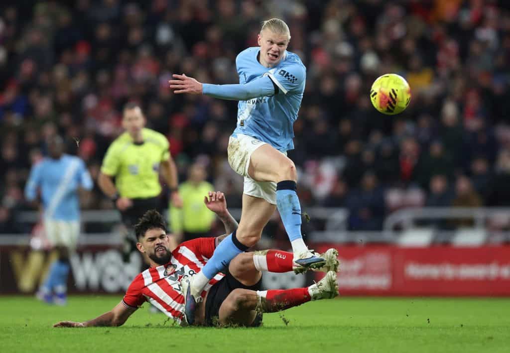 An intense soccer match featuring a player in blue jumping over an opponent in red and white during a competitive game. The stadium crowd is visible in the background, highlighting the energy and action on the field.