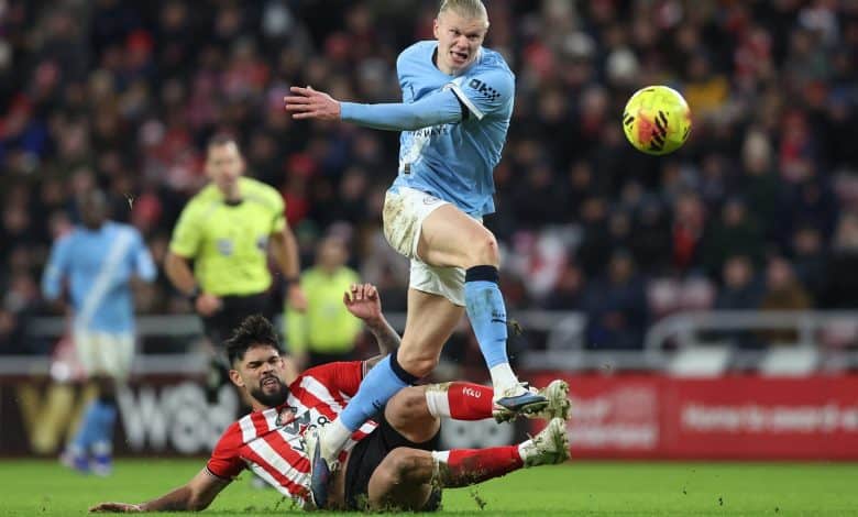 An intense soccer match featuring a player in blue jumping over an opponent in red and white during a competitive game. The stadium crowd is visible in the background, highlighting the energy and action on the field.