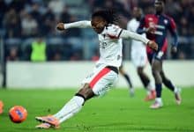 Soccer player in action, kicking the ball on the field with opposing players in the background.