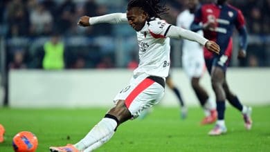 Soccer player in action, kicking the ball on the field with opposing players in the background.