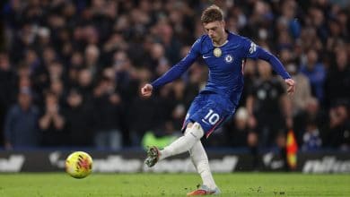 Soccer player in blue jersey kicking football in match, demonstrating skill and agility on the field, with a vibrant crowd watching in the background.