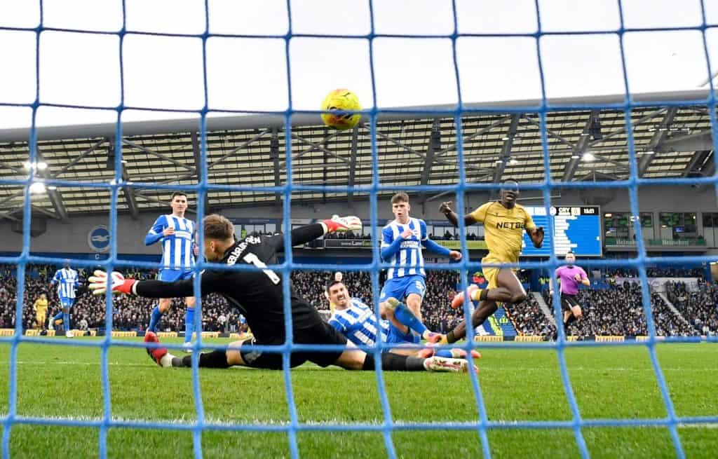Exciting moment of a soccer game with players in blue and white uniforms, goalkeeper diving to save the shot, and the ball heading towards the net.