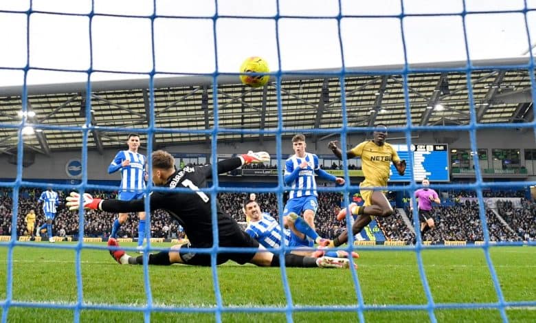 Exciting moment of a soccer game with players in blue and white uniforms, goalkeeper diving to save the shot, and the ball heading towards the net.